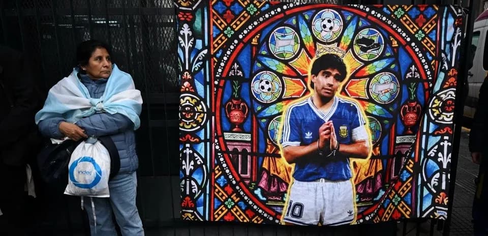 Ayol Argentina bayrog'ini o'rab olgan, devorda Diego Maradona rasmli plakat