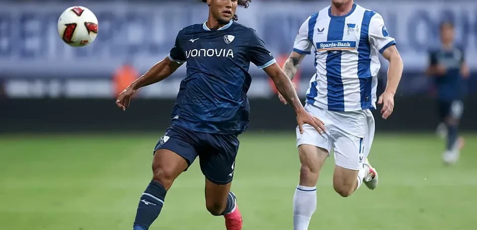 Player in dark kit chasing ball with opponent behind