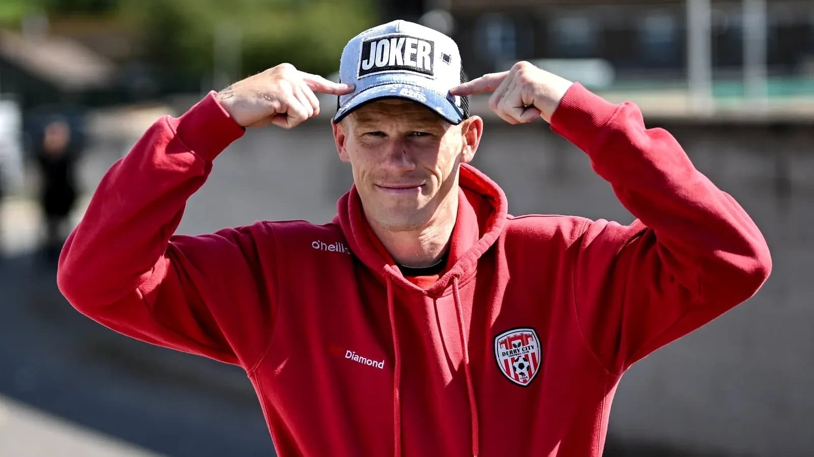 Shirted man pointing at 'JOKER' cap outdoors Shirted man pointing at 'JOKER' cap outdoors